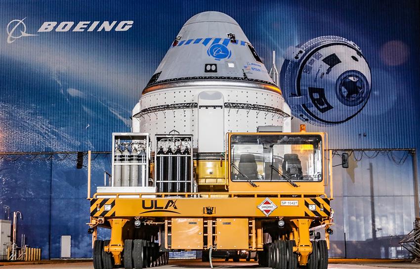 A spacecraft is being moved on a motorized transporter in the Boeing integration hall, where a mural shows the logo.