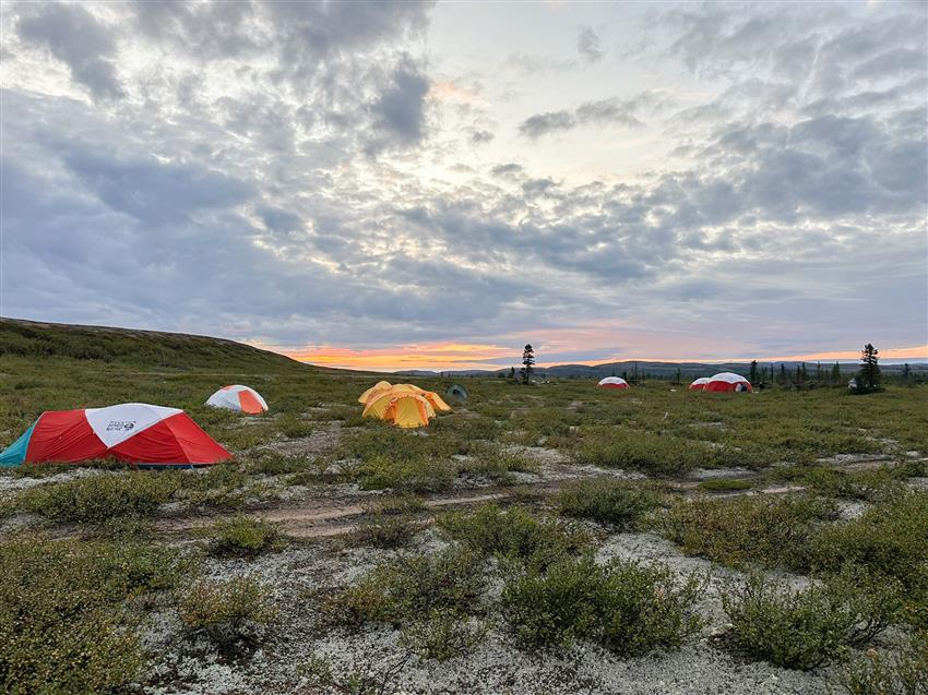 Several camping tents spread out in the tundra with the setting sun in the distance.