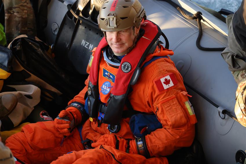 Jeremy est assis dans un bateau pneumatique. Sa combinaison spatiale arbore à l'épaule un écusson du drapeau du Canada.