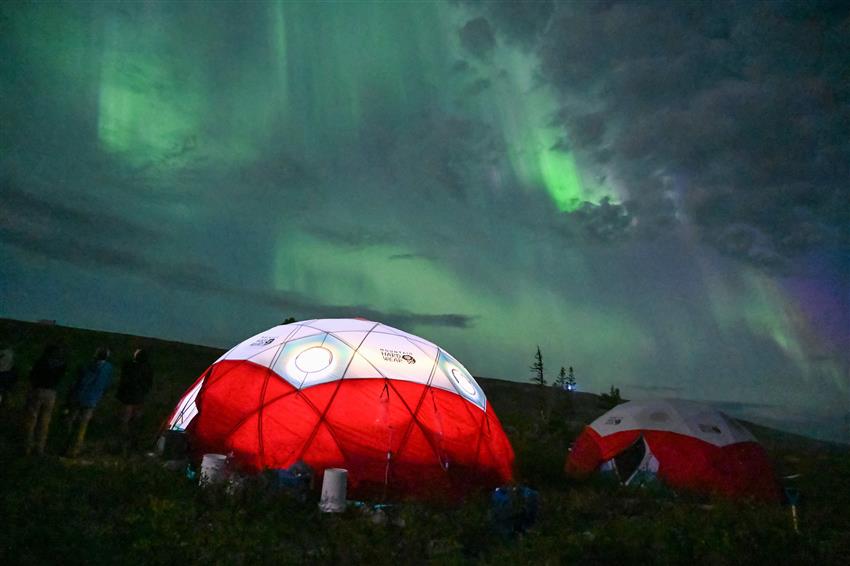 Green northern lights over two large camping tents. The tent in the foreground is illuminated.